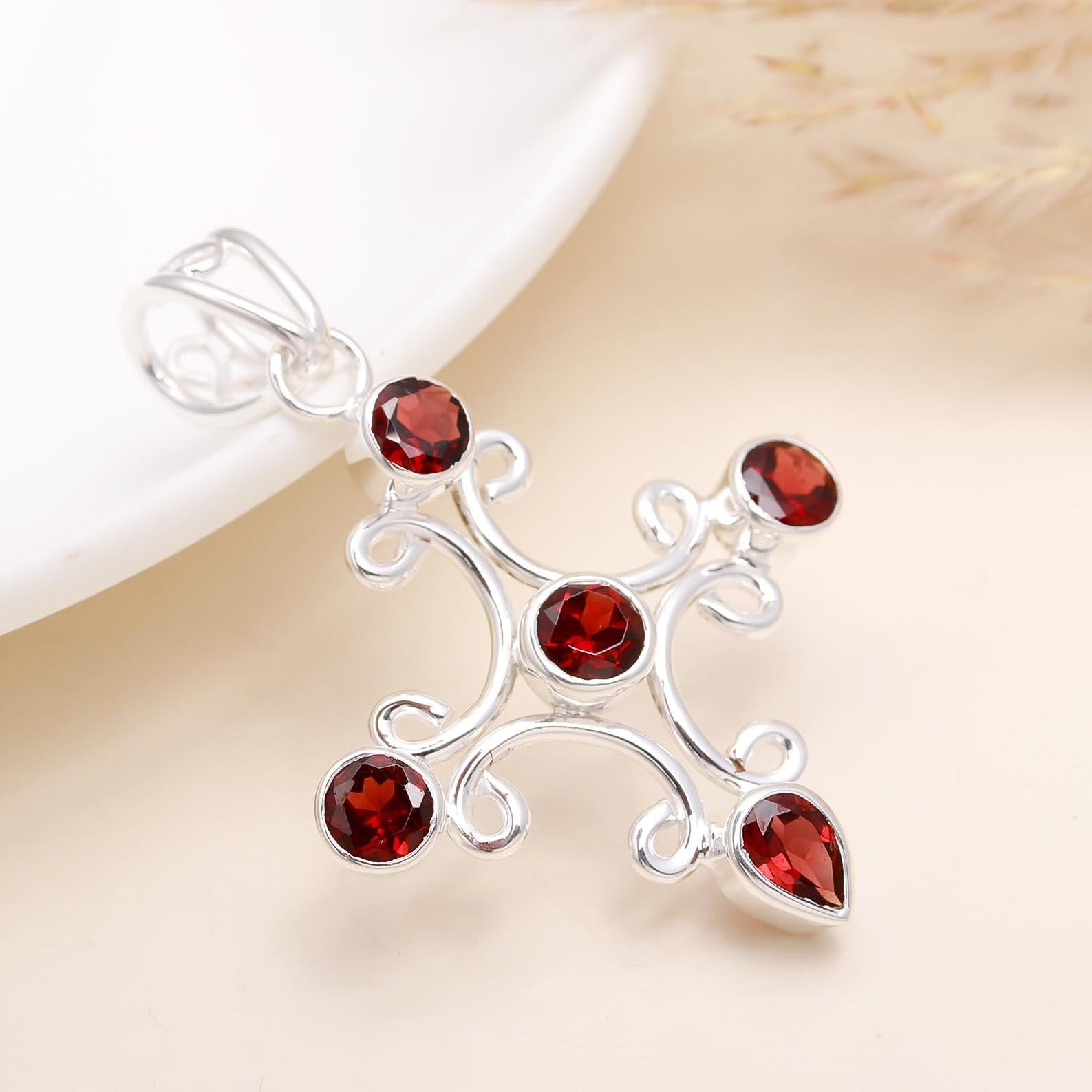 A sterling silver cross-shaped pendant with a central garnet gemstone, surrounded by smaller clear crystals, displayed on a white plate with dried grass in the background.