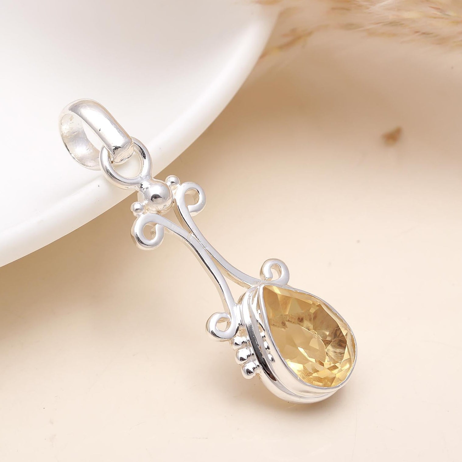 A 925 sterling silver citrine pendant with a decorative design, placed on a white surface next to a dried plant, showcasing its silver finish and the citrine gemstone inside.