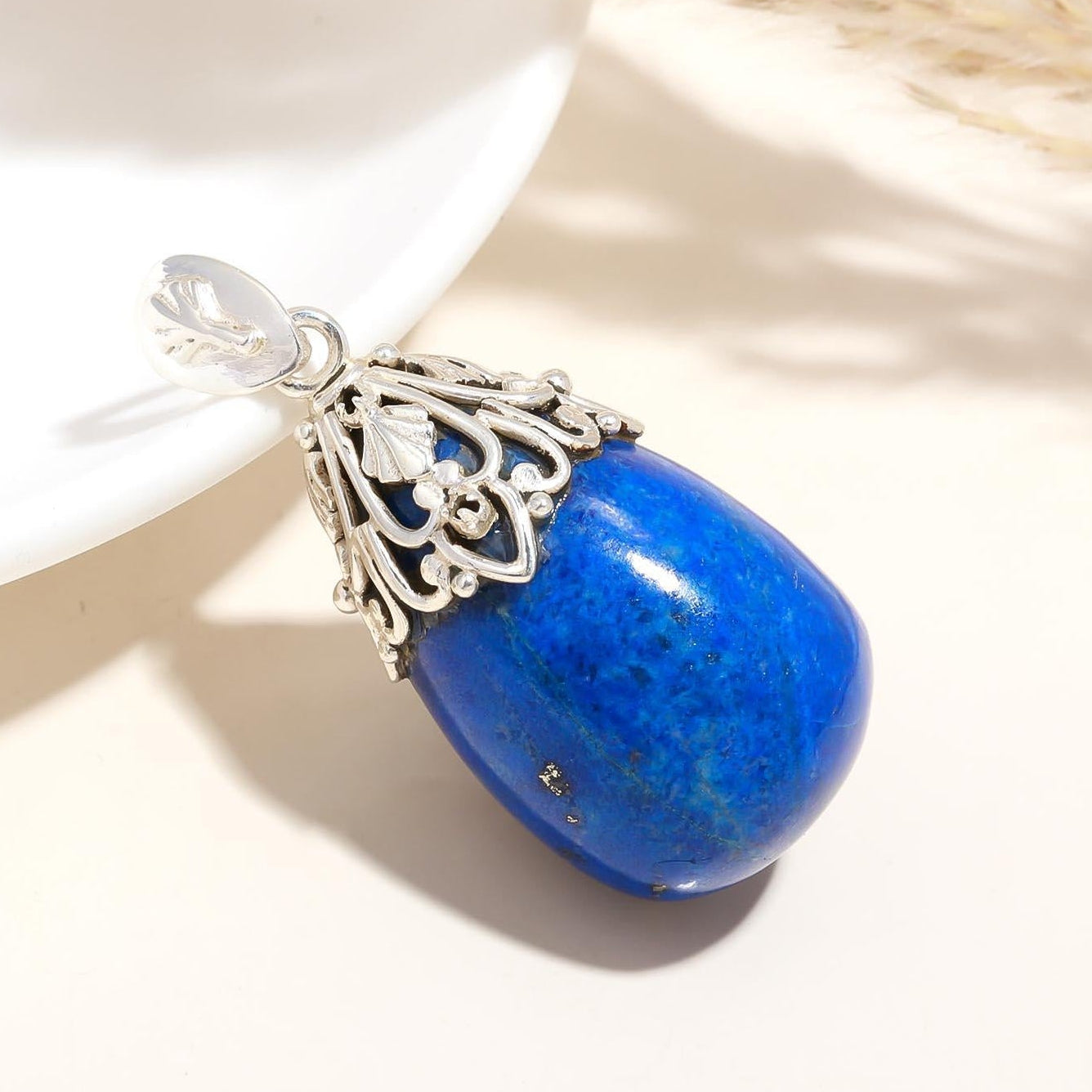 Blue stone pendant with silver decorative element on a white surface with dried plants.