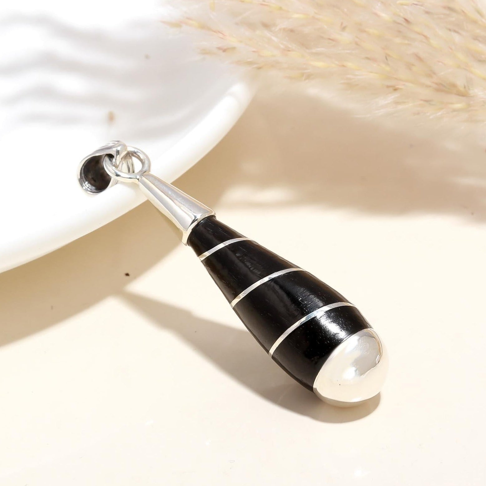A black and white strip design sterling silver pendant on a white surface, with a white plate and some dry plants in the background.