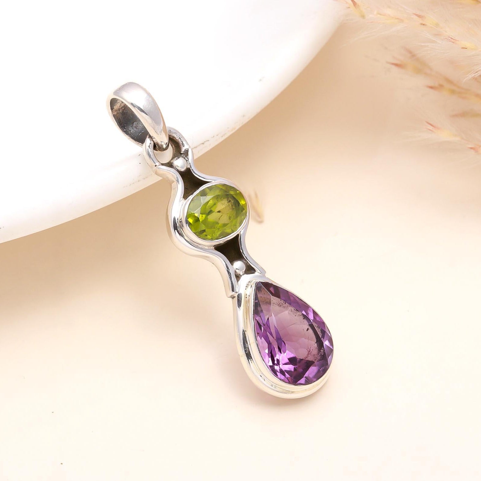 A sterling silver pendant featuring a drop design with one round amethyst gemstone on top and one teardrop peridot gemstone below, set against a neutral background.