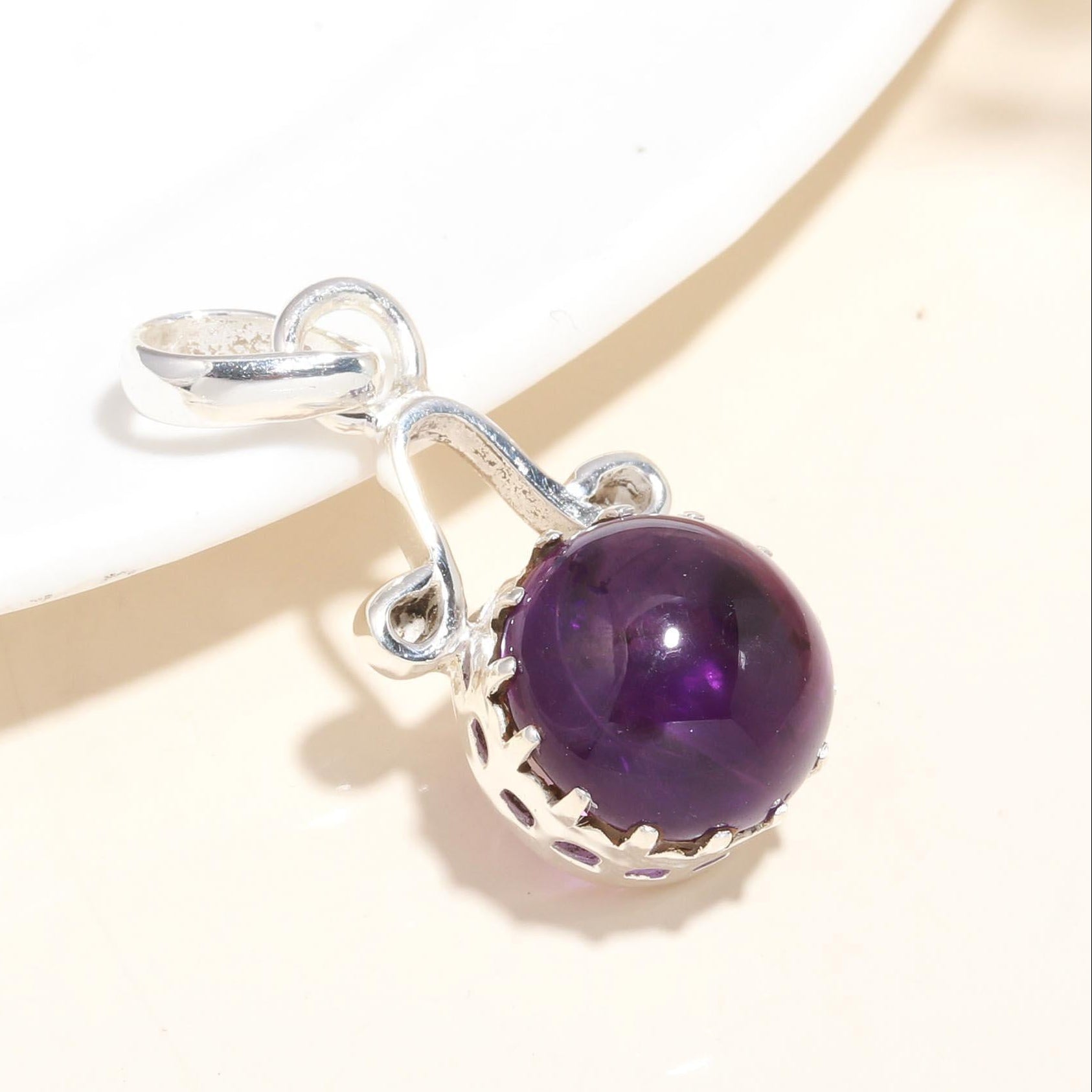 A polished purple amethyst gemstone set in a sterling silver sphere-shaped pendant, displayed against a beige background.