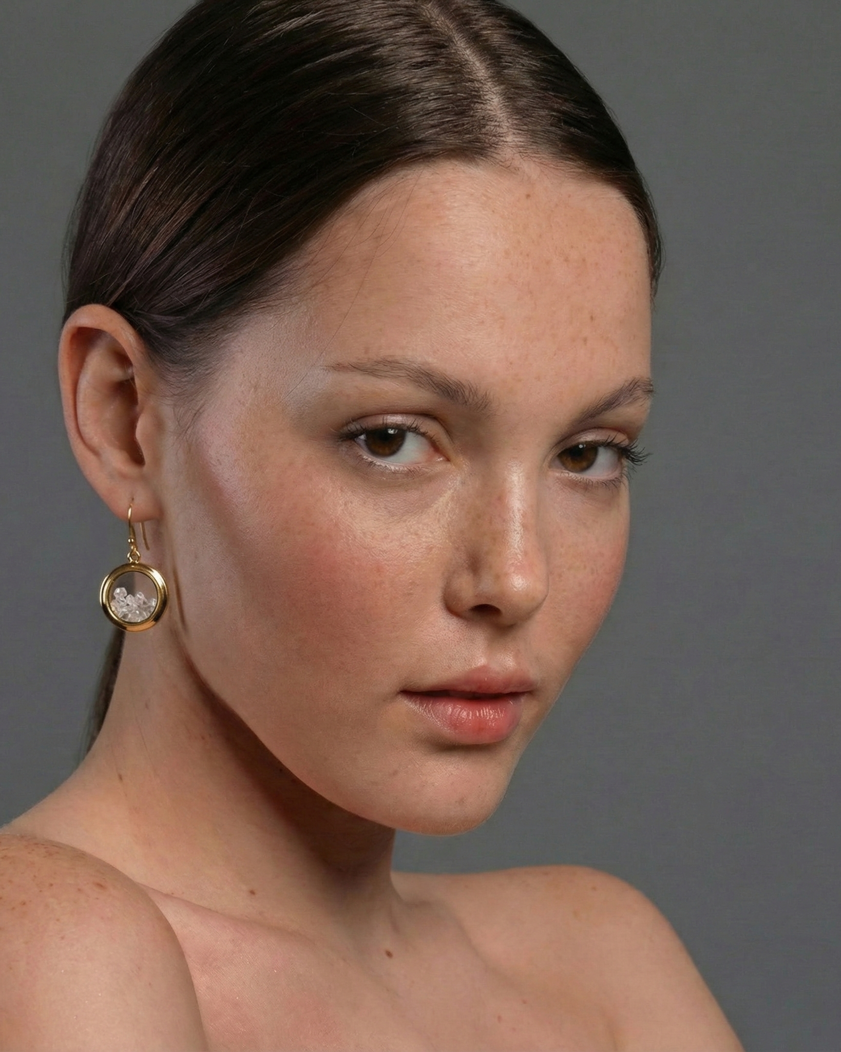 Close-up of a woman wearing a gold earring with a clear gemstone against a gray background