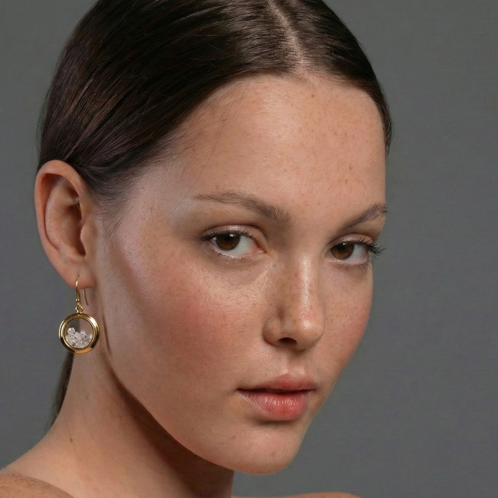 Close-up of a woman wearing a gold earring with a clear gemstone against a gray background