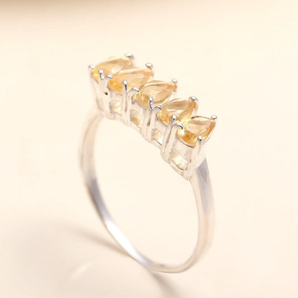 A silver ring featuring five pear-cut yellow gemstones set in a row, displayed on a soft beige background.