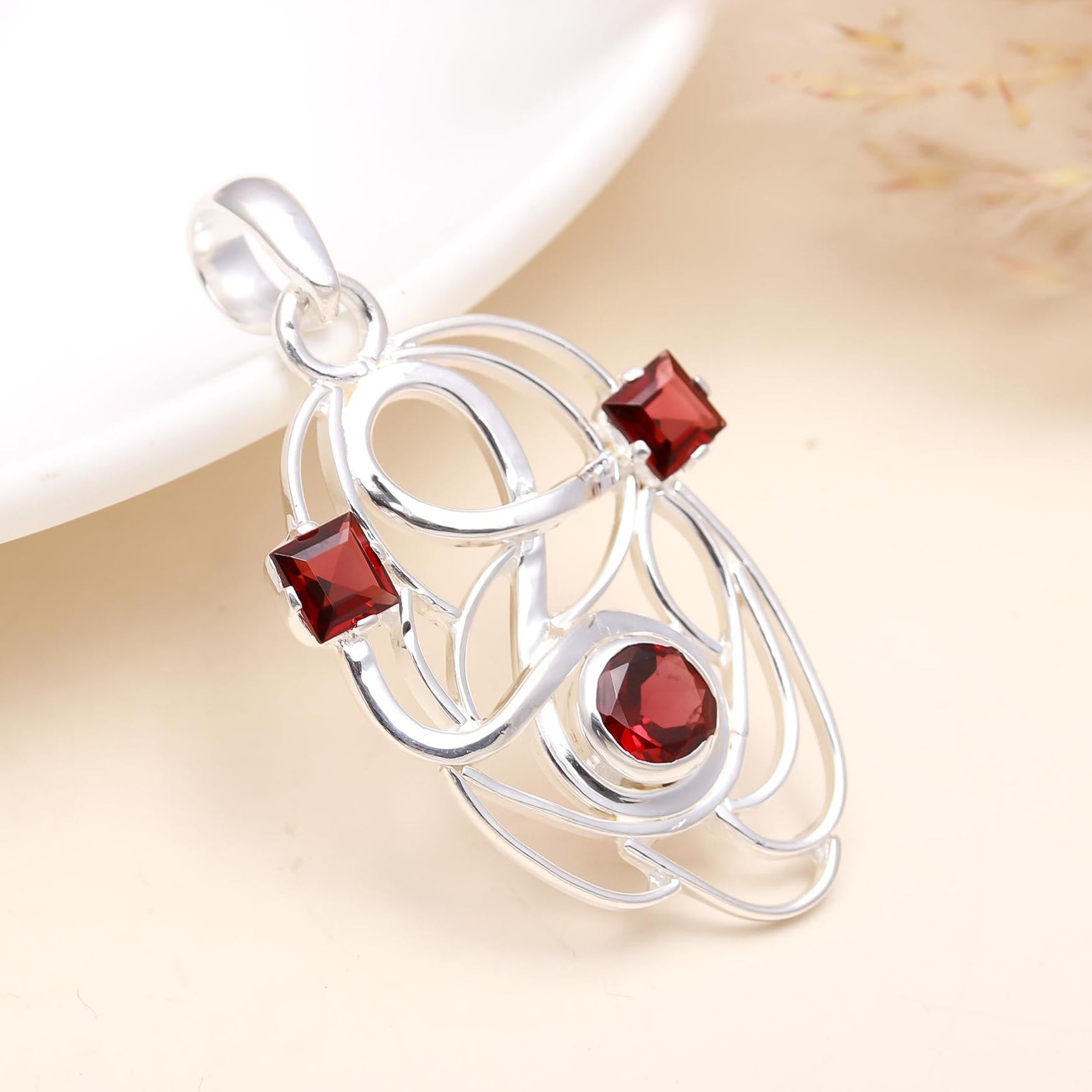 A fine sterling silver pendant with a natural garnet gemstone in a minimalist design, displayed on a white plate with dry grass in the background.