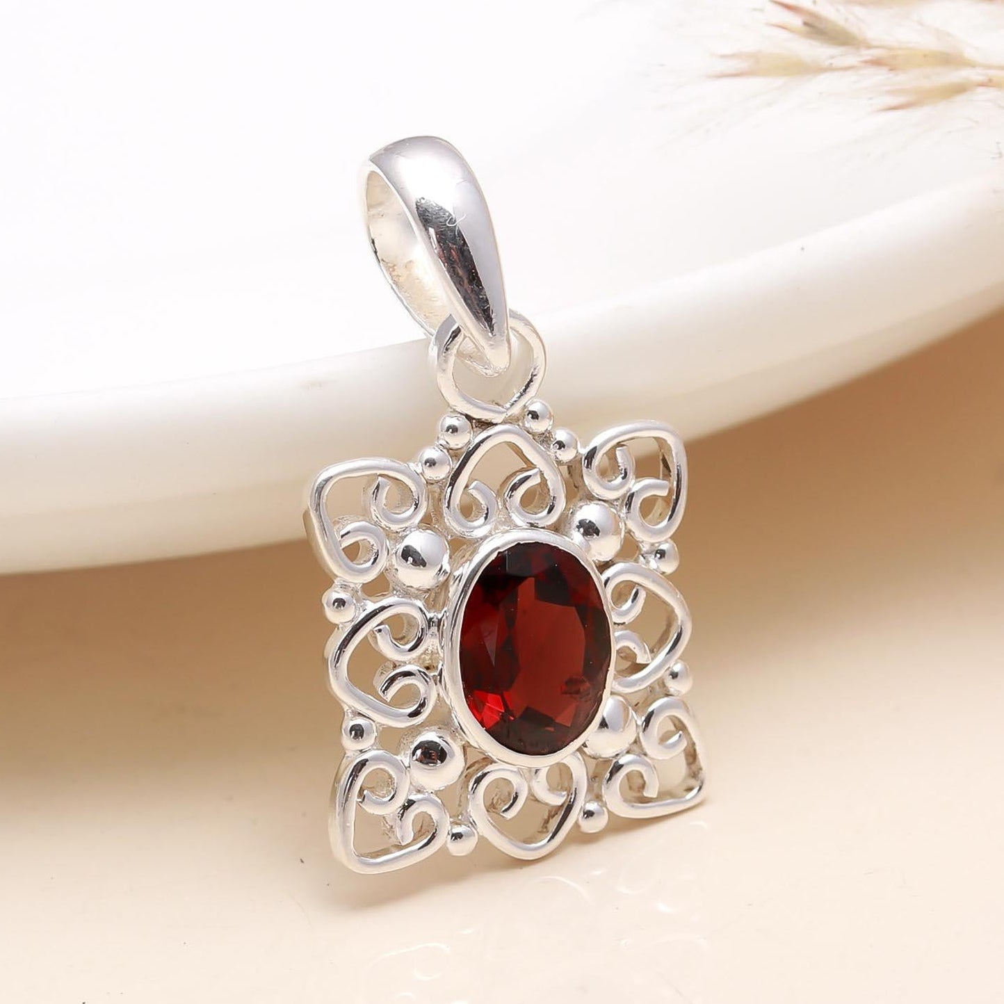 A square-shaped garnet gemstone pendant with a silver metallic setting, displayed on a white plate with a wired silver pouch in the background.