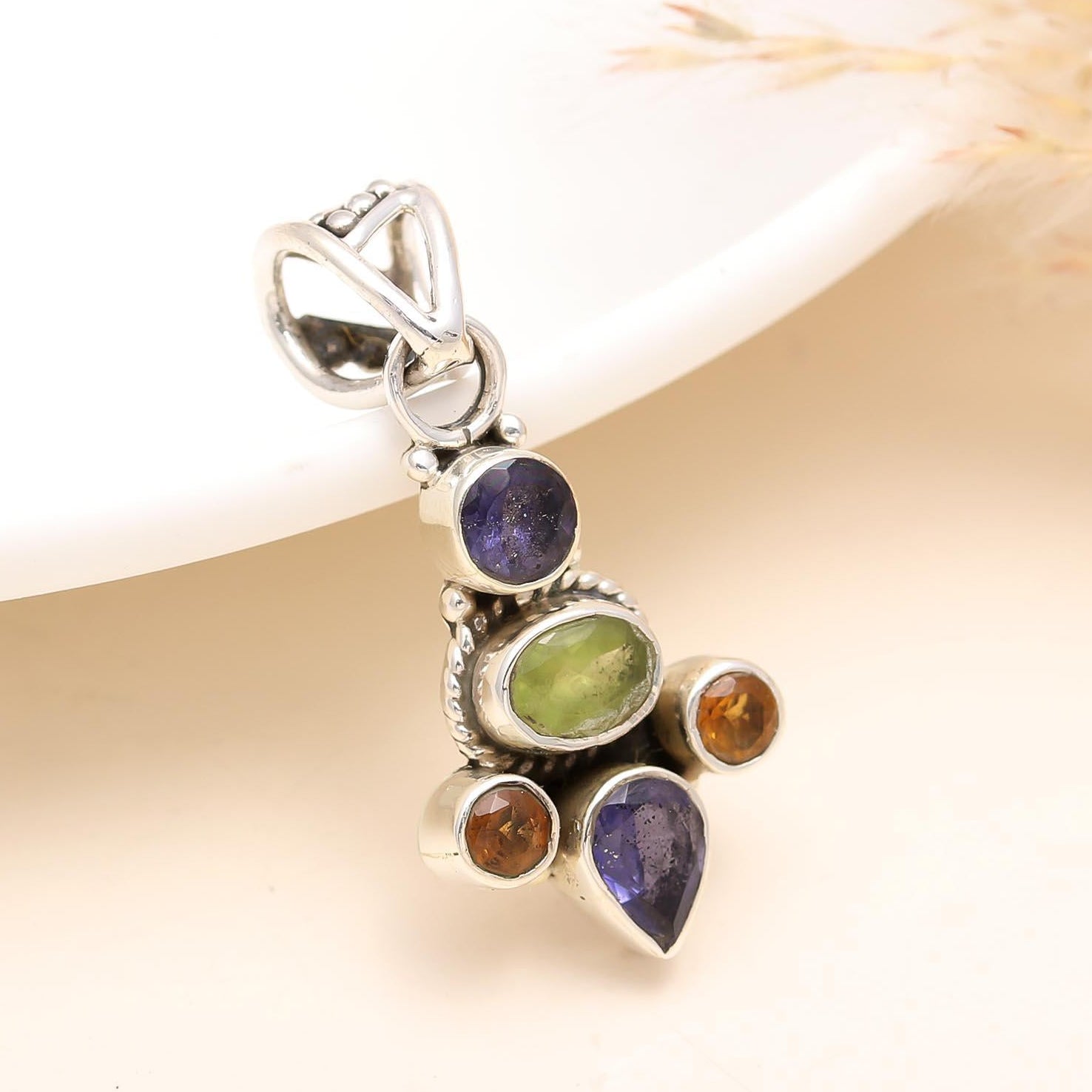 A sterling silver pendant featuring a multi-colored gemstone design, displayed against a neutral background with dried plant material in the foreground.