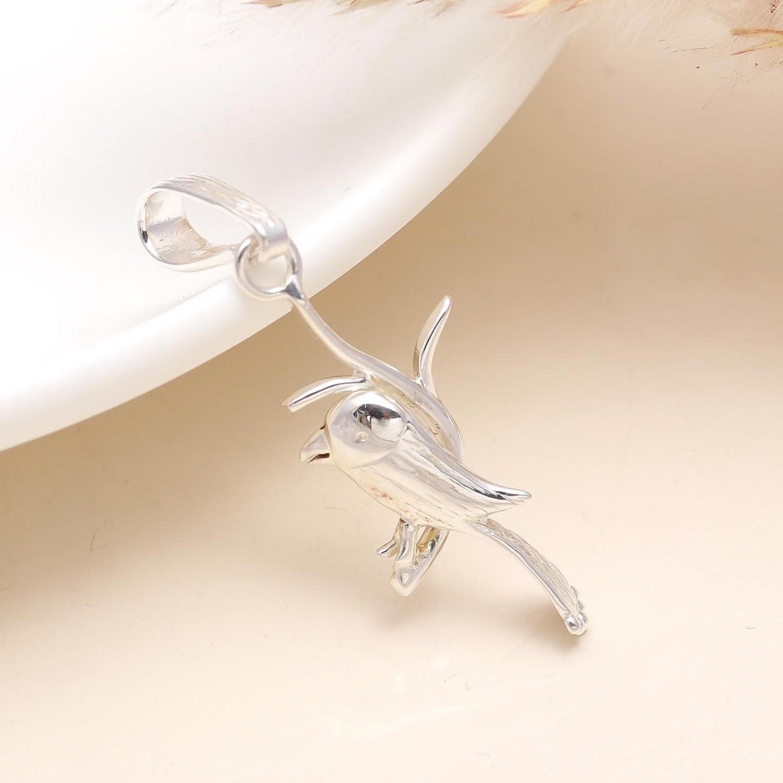 A sterling silver bird-shaped pendant on a chain, displayed over a white plate with a feather in the background.