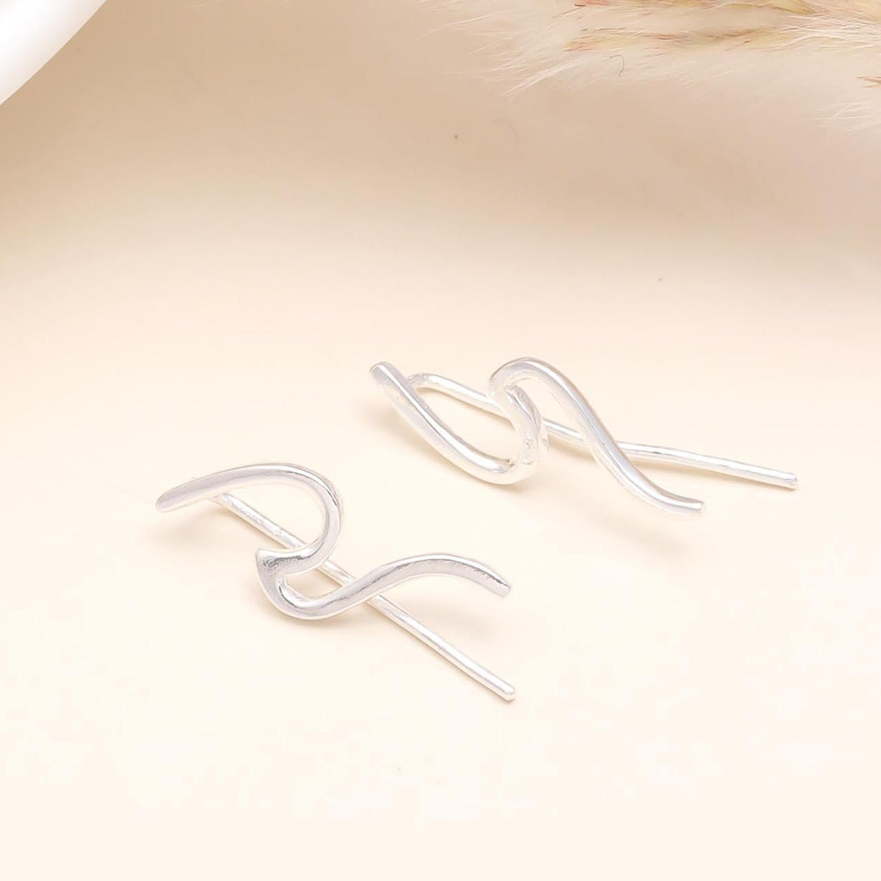 A pair of 925 sterling silver wave ear climber earrings positioned on a beige surface, with a white object and a dried plant in the background.