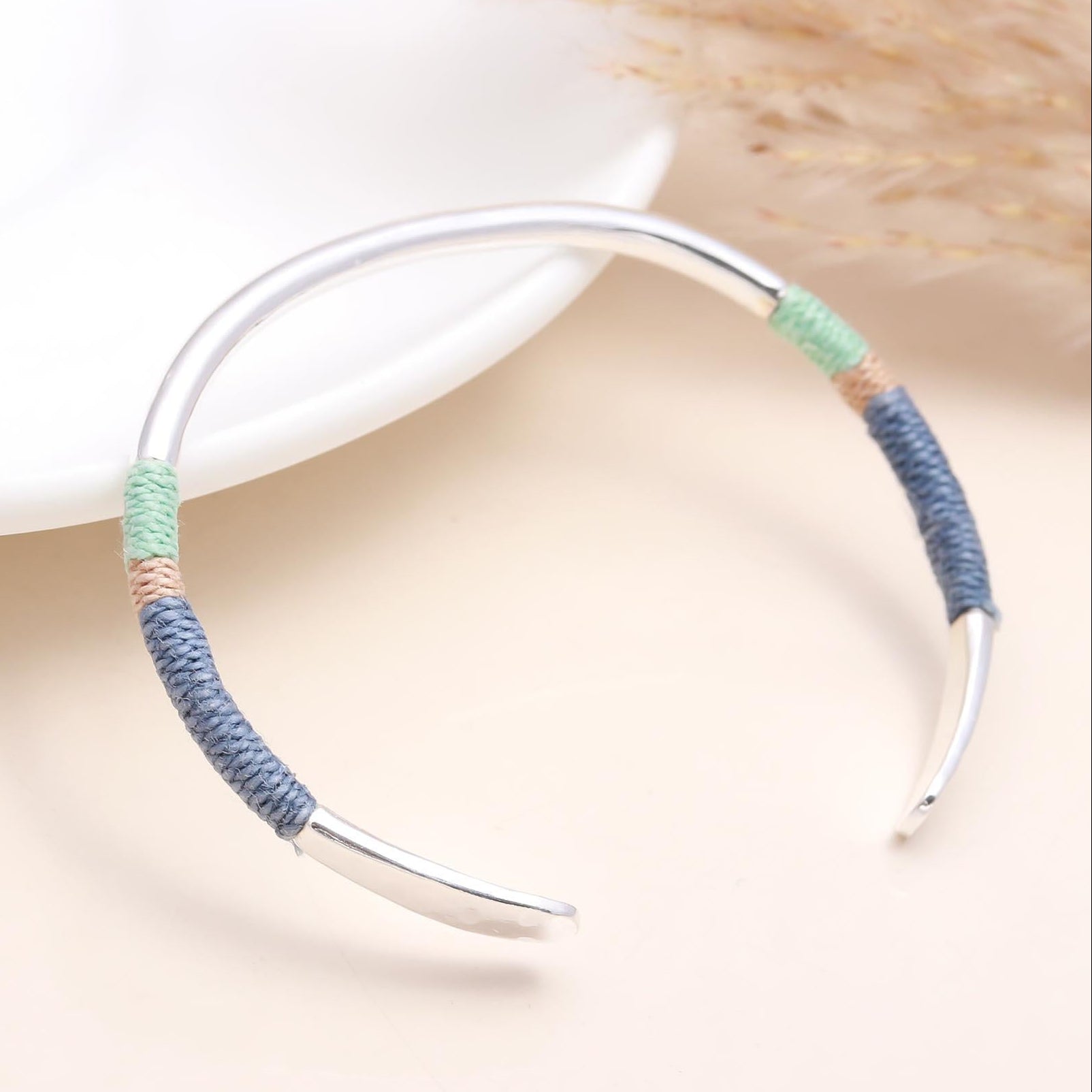 A 925 sterling silver cuff bracelet with thread-wrapped details, displayed on a white surface with a dried plant in the background.