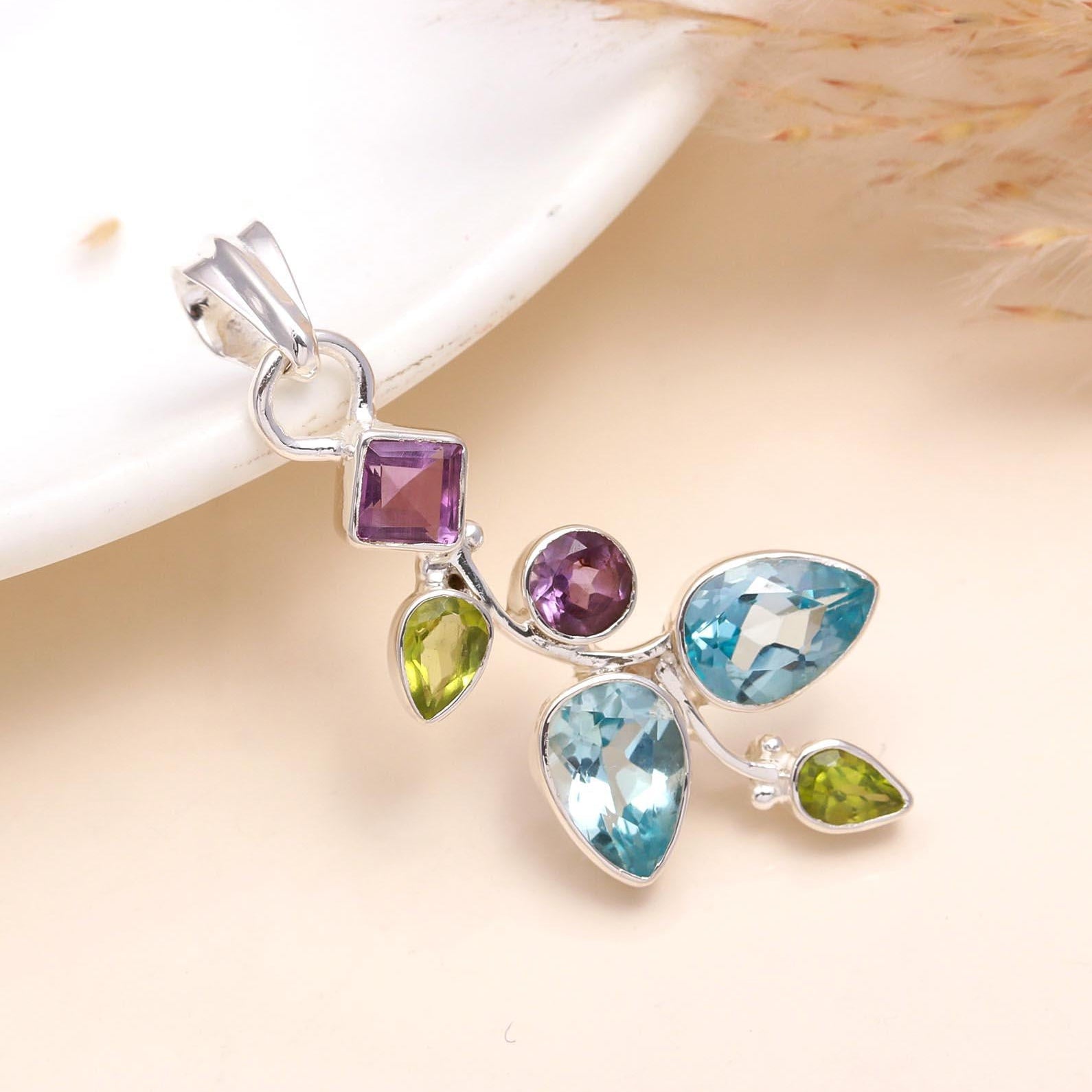 A sterling silver pendant featuring a multi-gemstone arrangement with blue topaz, amethyst, and peridot, presented on a white surface.