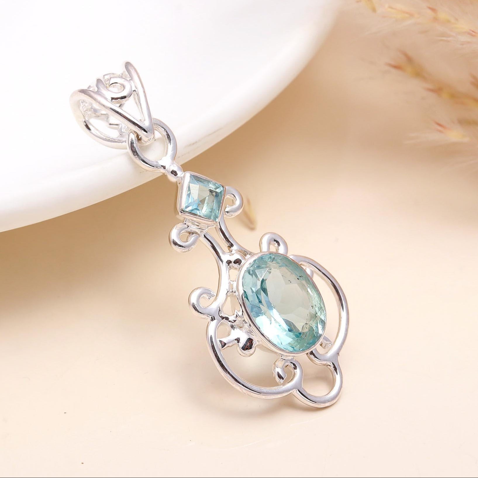 A vintage-style sterling silver filigree pendant featuring a central blue topaz gemstone, accompanied by smaller blue stones, presented on a white surface.
