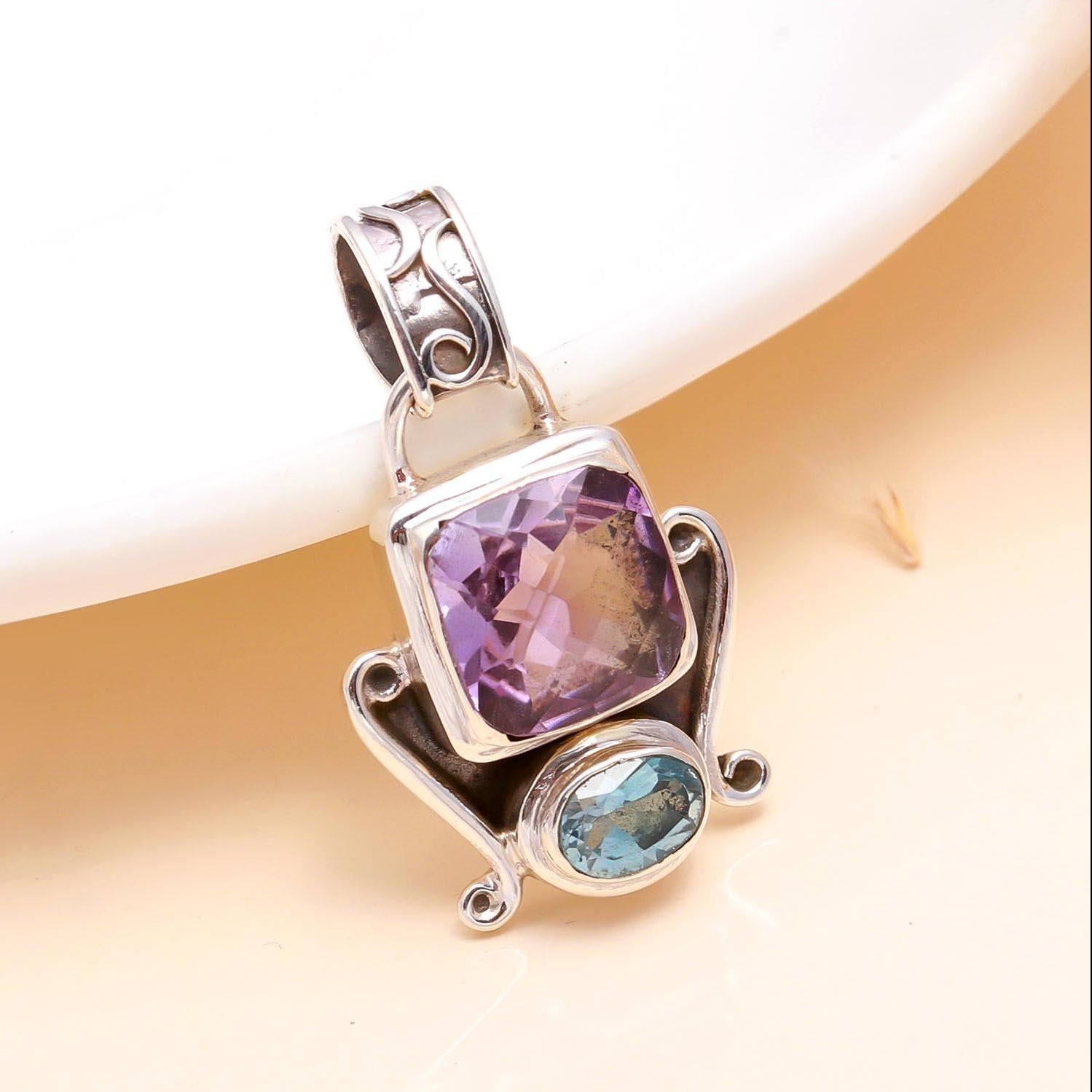 A sterling silver pendant featuring a large amethyst gemstone with a smaller blue topaz gemstone to the side, set against a neutral background.