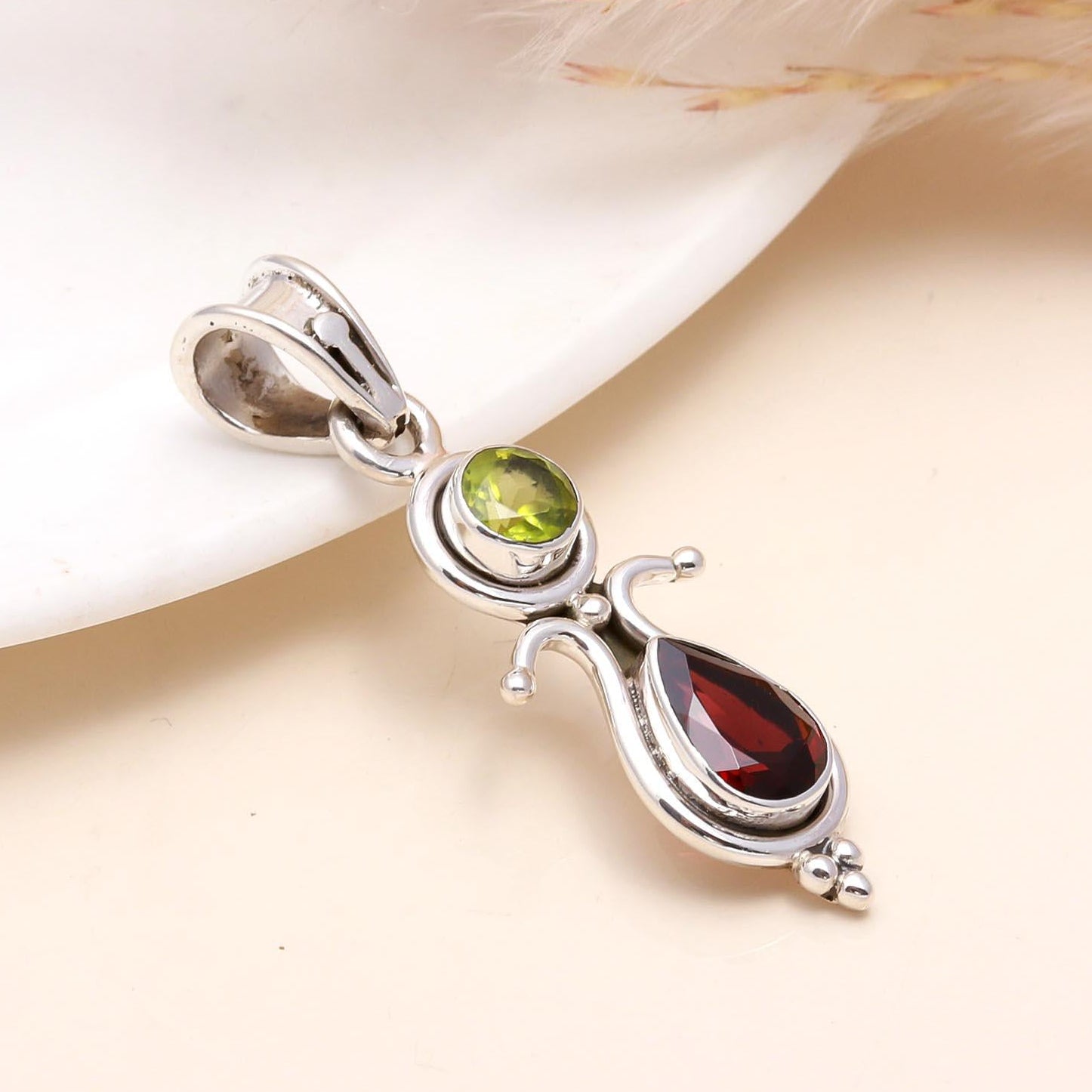 A sterling silver pendant featuring a vintage style design with one garnet and one peridot gemstone, displayed against a neutral background.