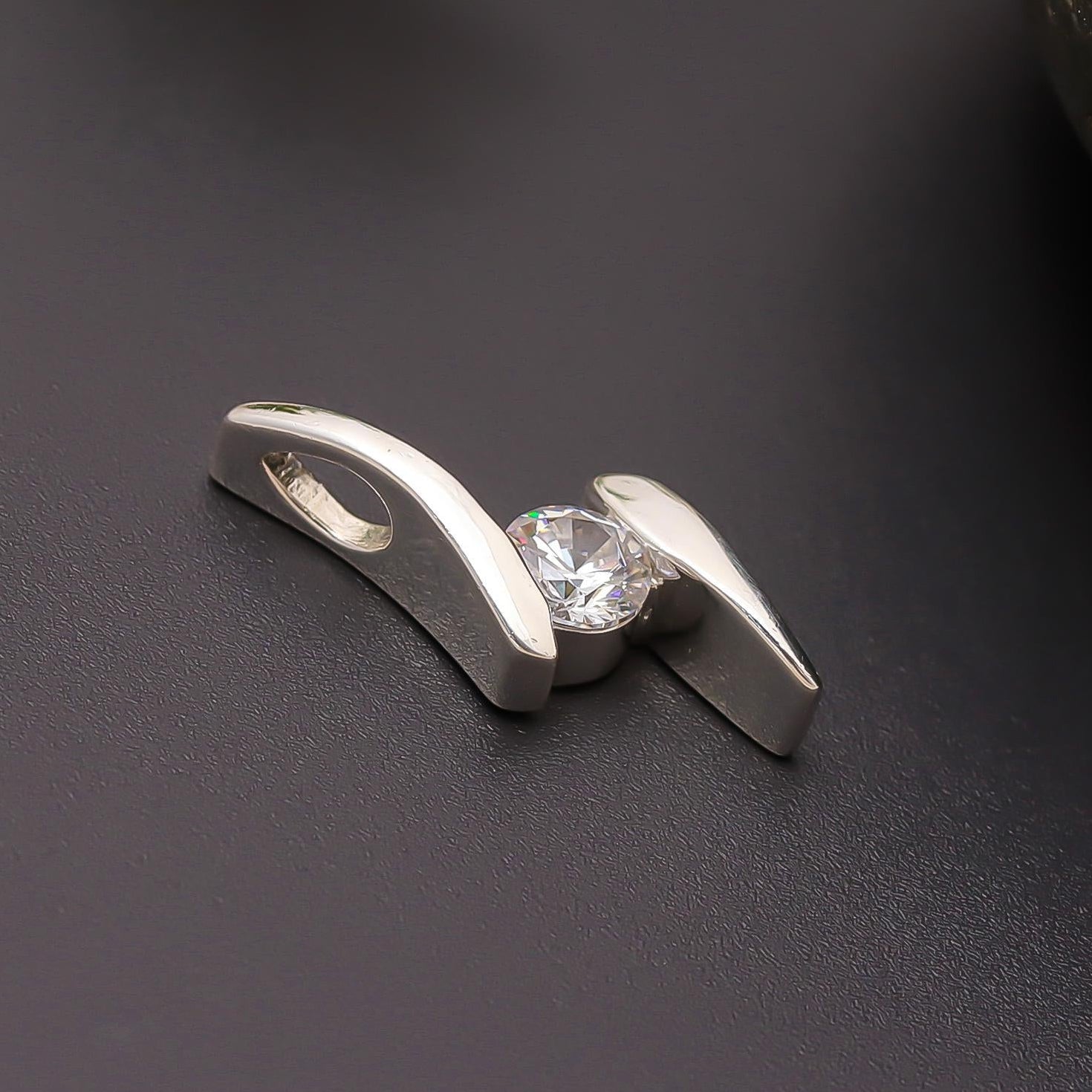 A sleek silver solitaire pendant with a round-cut clear stone, displayed against a dark background with a rock and a green leaf in the background.
