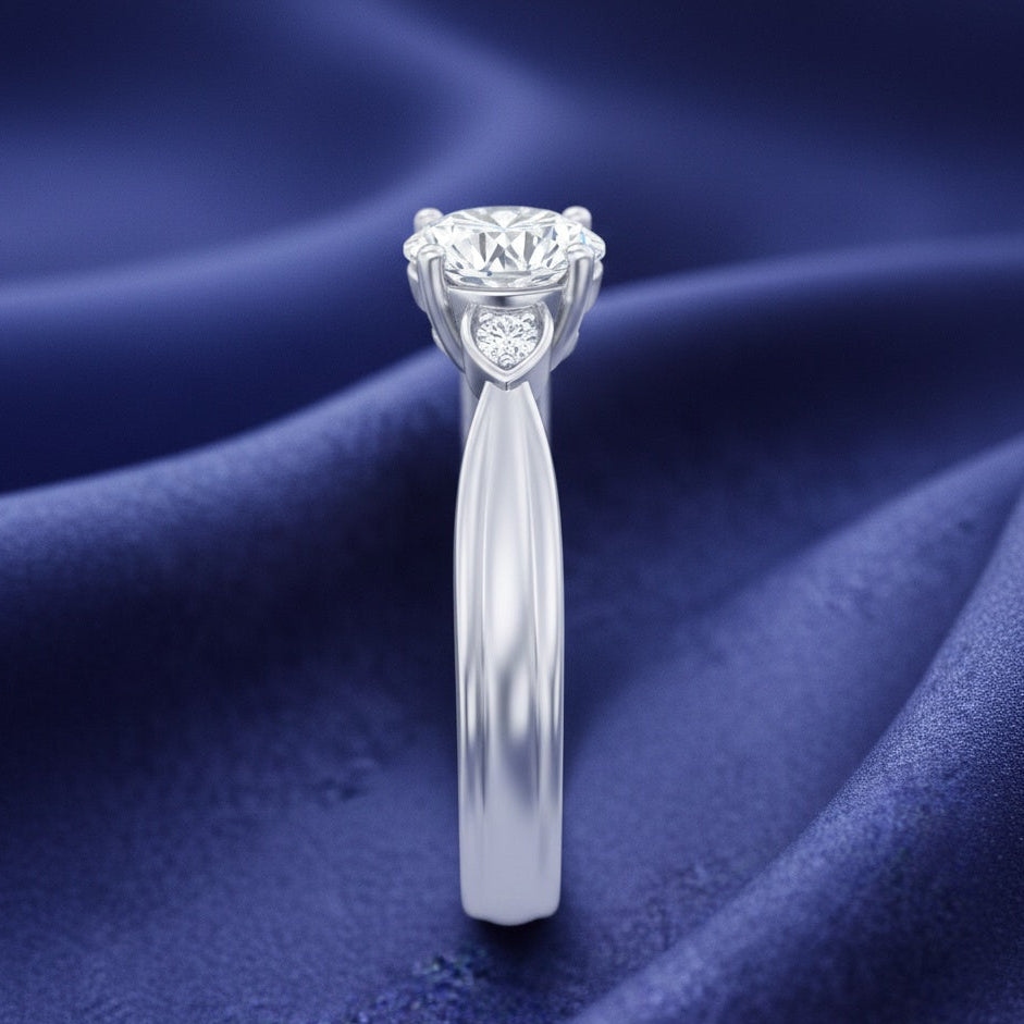A silver engagement ring with a heart-shaped diamond halo and a round center stone, displayed on a draped dark blue satin fabric background.