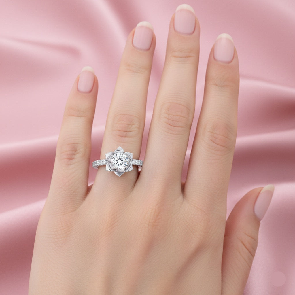 A silver-toned diamond ring with a floral rose-cut center stone and small side diamonds, shown worn on a hand against a soft pink satin background.